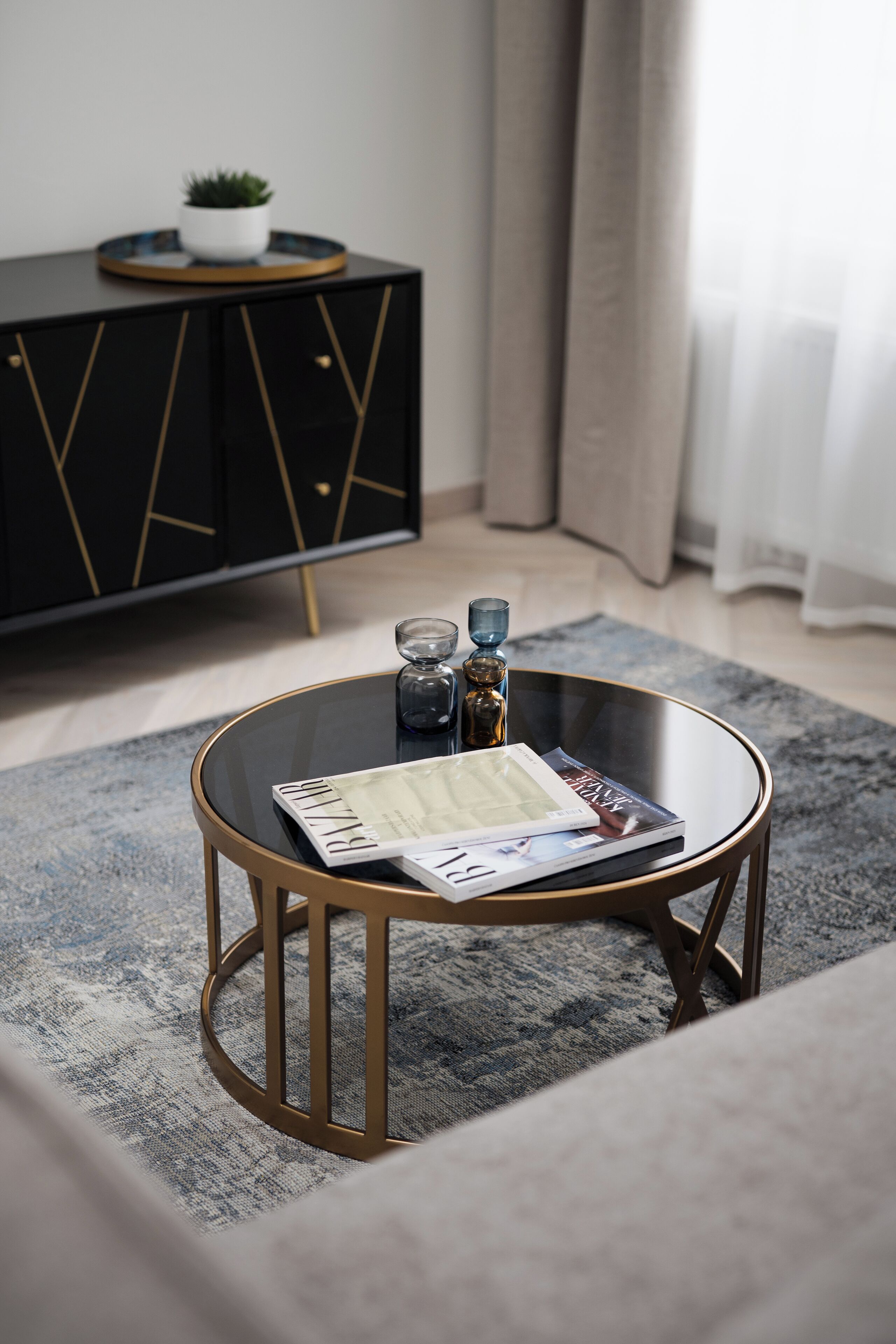 A round glass coffee table with a gold frame, a magazine, and a glass vase on a grey rug.