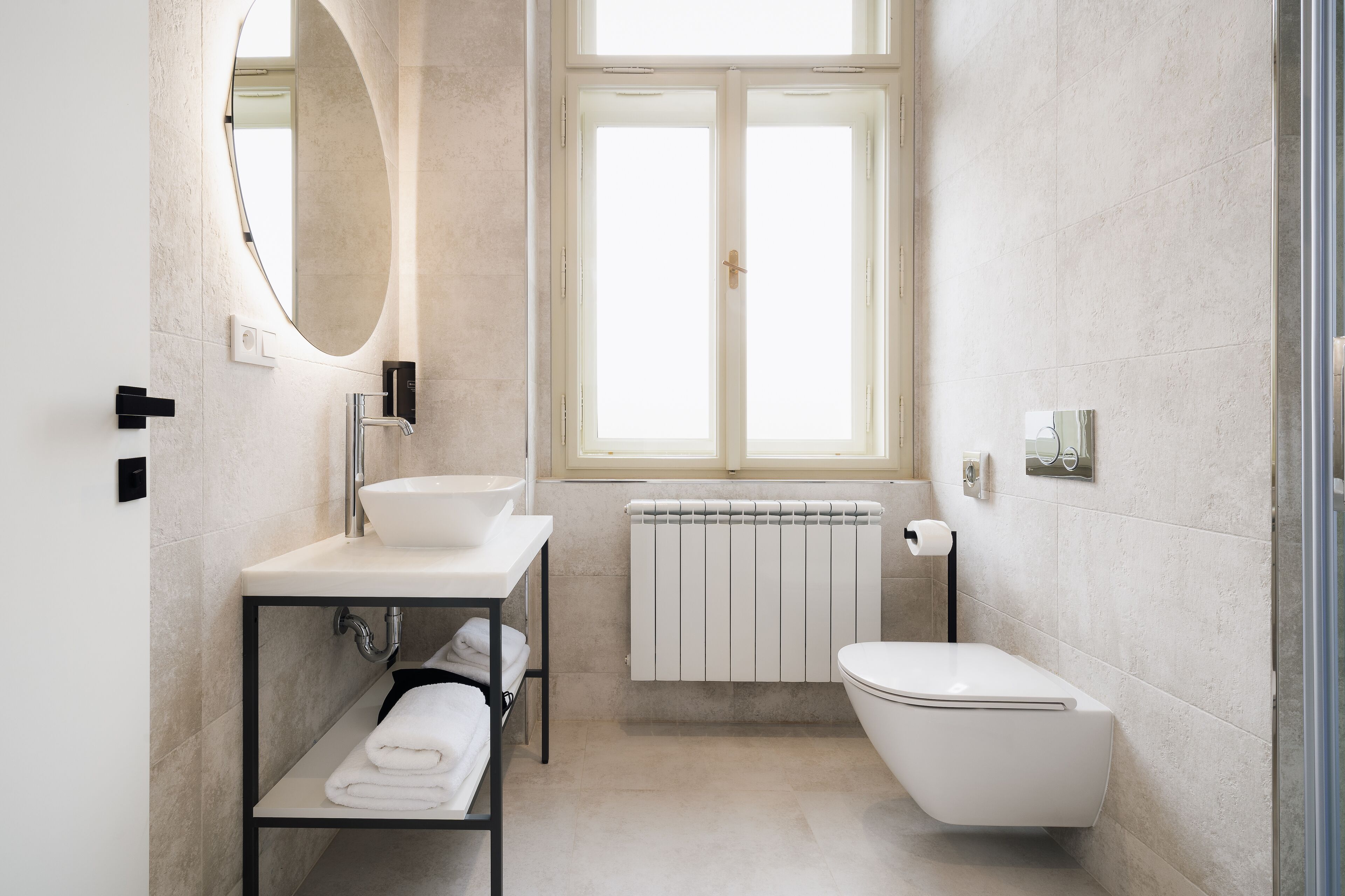Un bagno moderno con un lavandino bianco, uno specchio rotondo, un water e una finestra vista esterna.