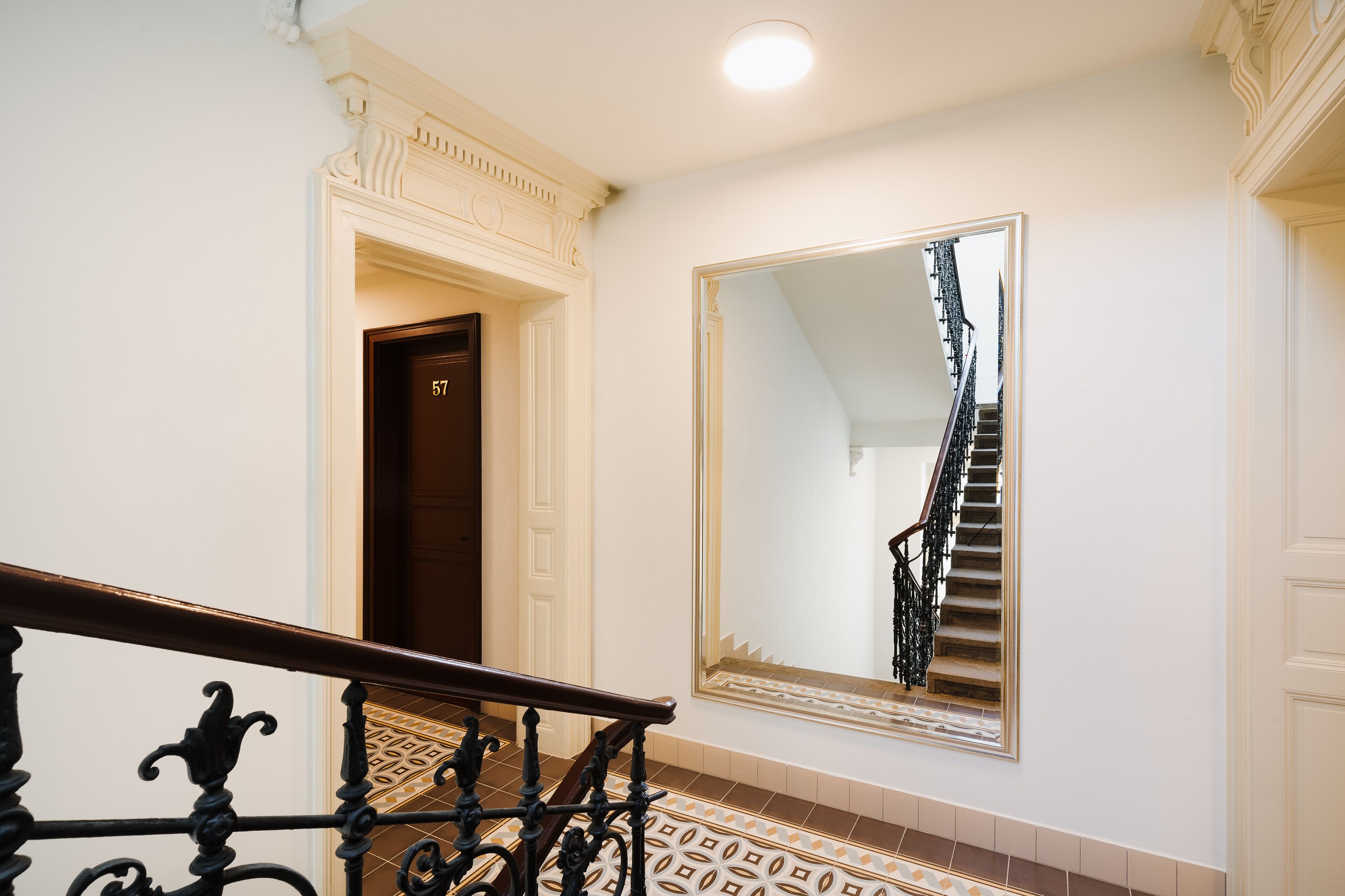 A staircase with a handrail, a large mirror reflecting the stairs, and a door labeled "57" in a hallway with patterned flooring.