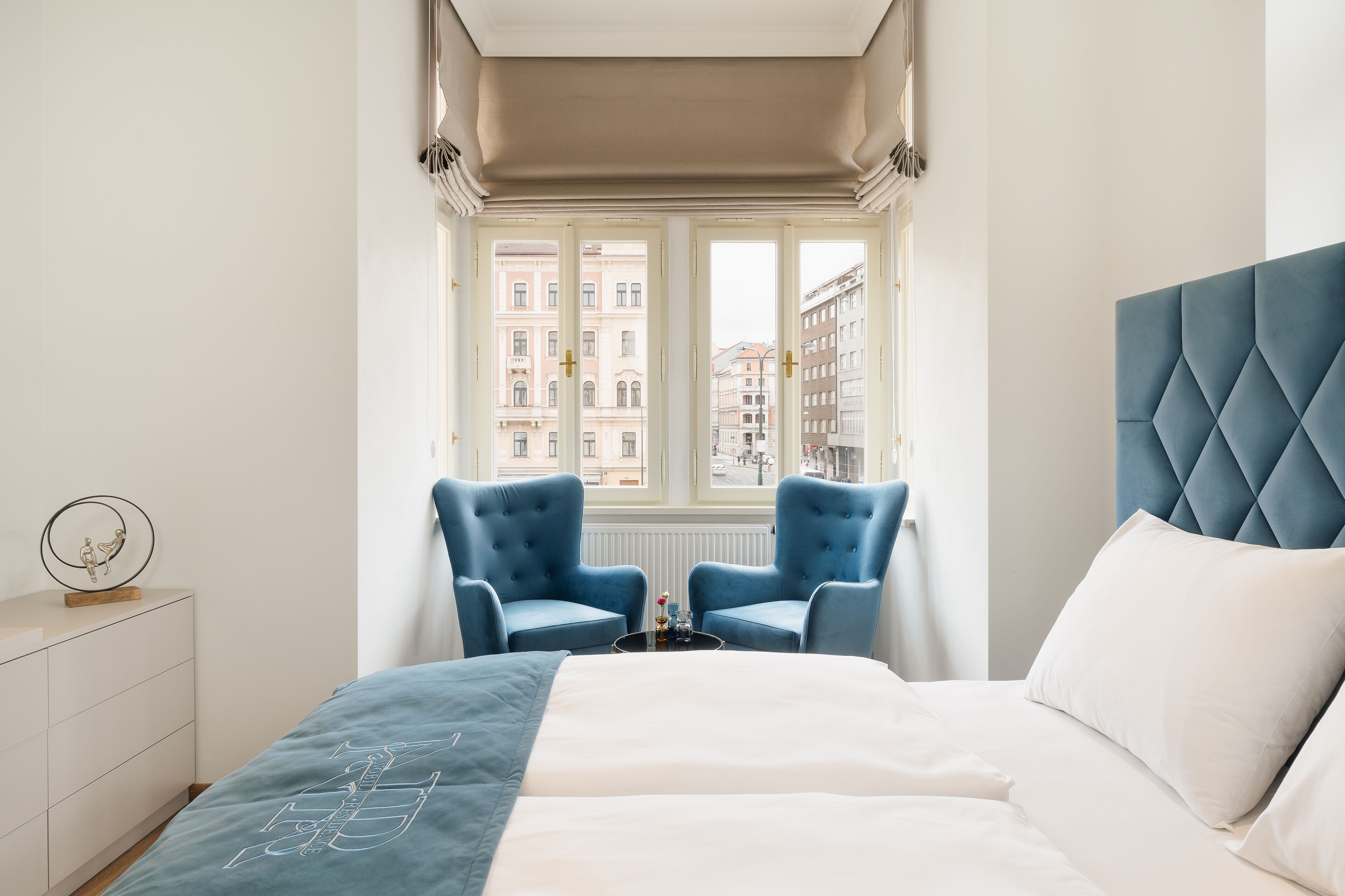 A hotel room with a bed, two blue armchairs, a small table with a lamp, and a window with a view of buildings.