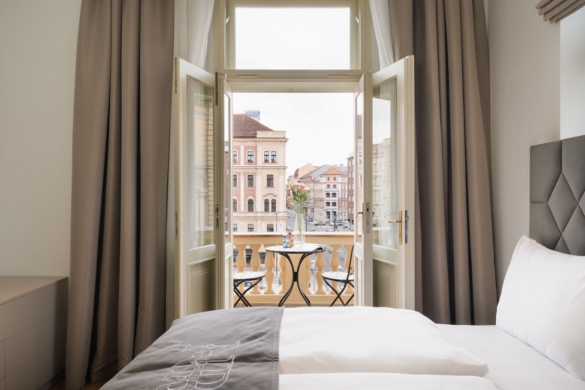 A hotel room with a bed, a small balcony with a table and chairs, and a view of buildings outside.