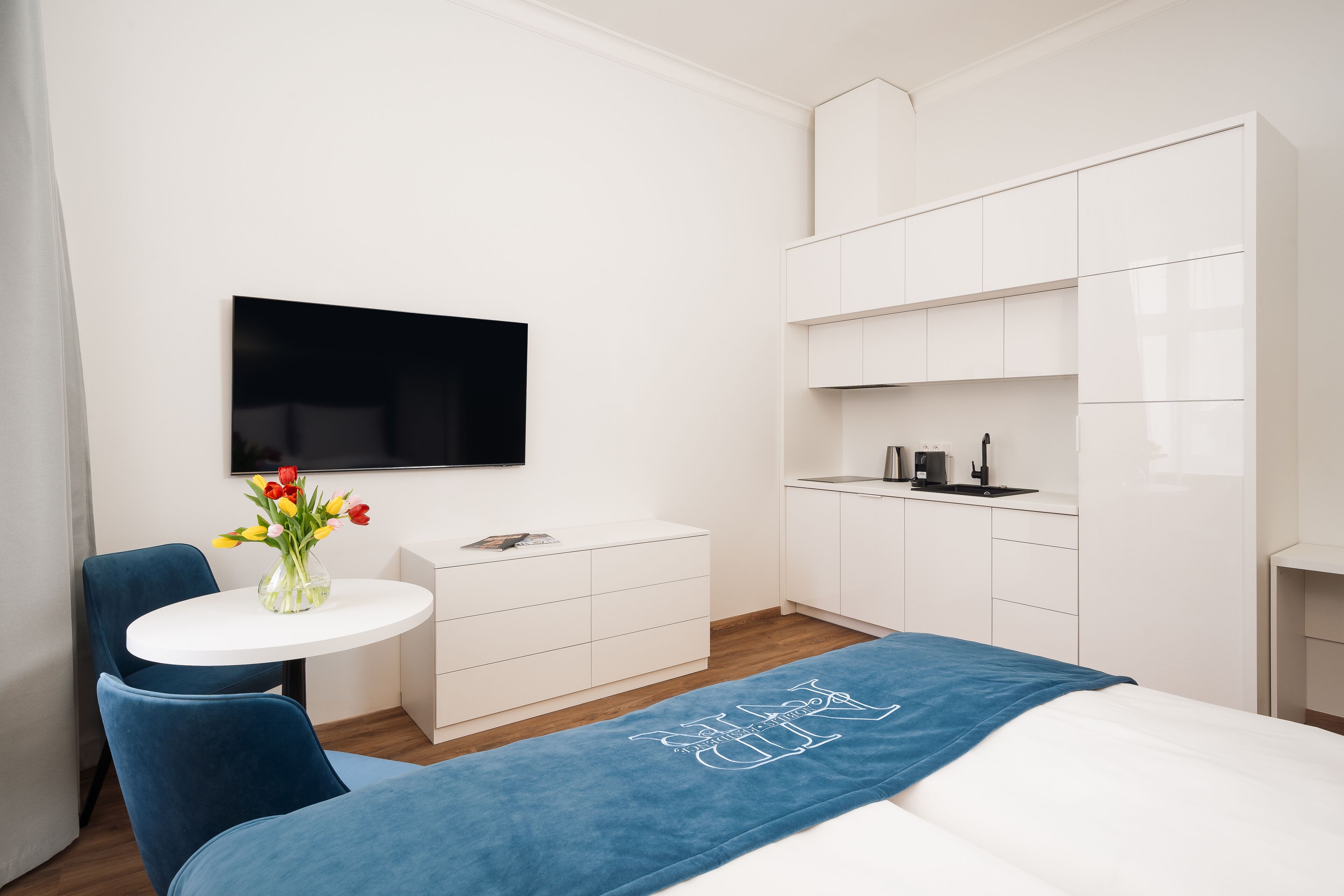 Een moderne hotelkamer met een bed, een klein rond tafeltje met een bloevas, een aan de muur bevestigde televisie en een witte kledingkast.