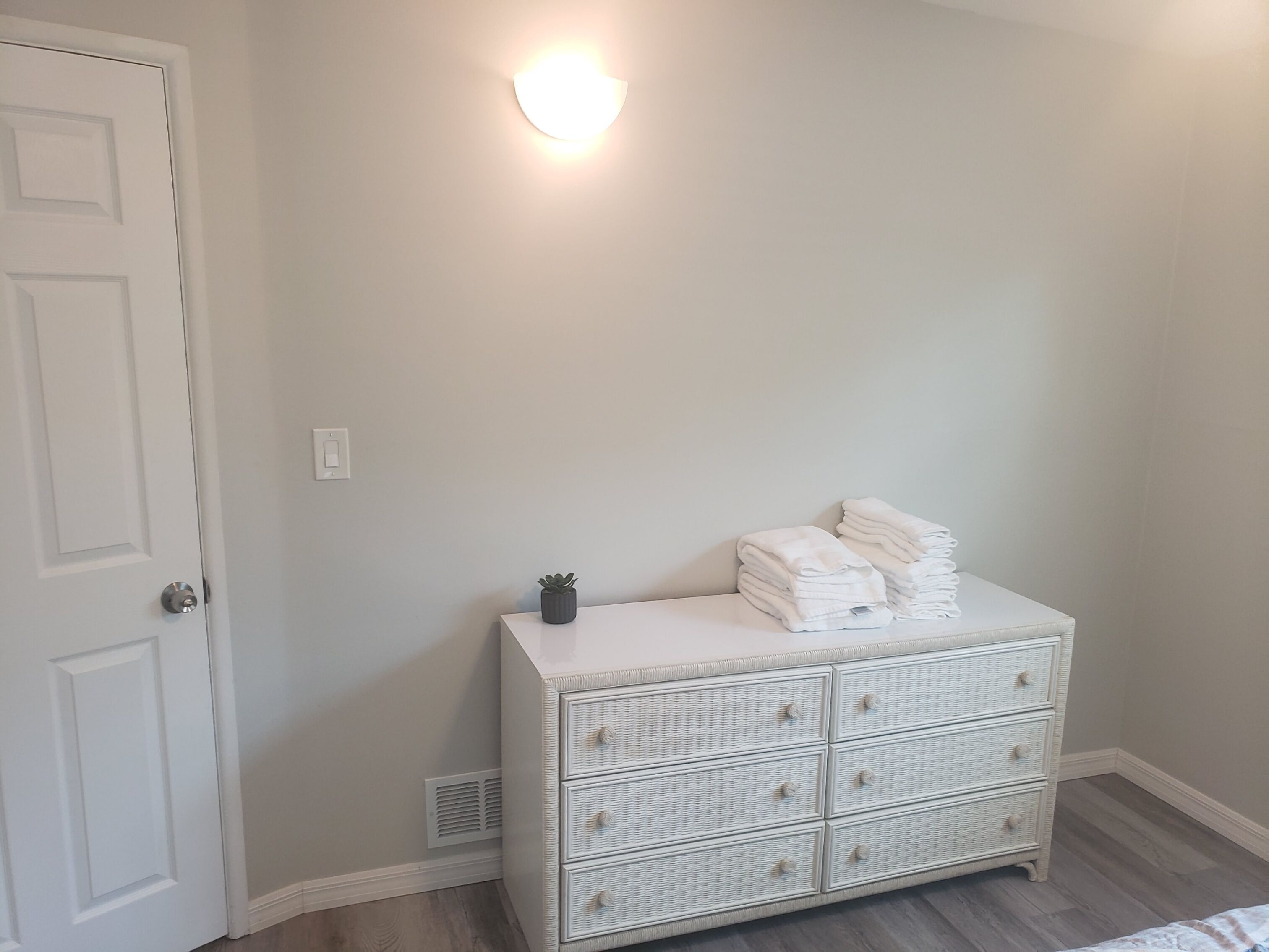 A room with a white door, a wall-mounted light, a white wicker dresser with drawers, and a small potted plant.