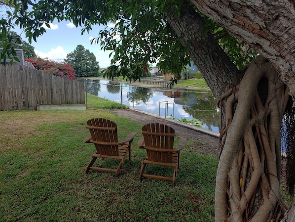 Two wooden Adirondack chairs facing a pond with a wooden fence and trees in the background.