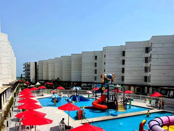 Una piscina con toboganes y zona de juegos acuáticos, rodeada de sombrillas rojas y sillas blancas.