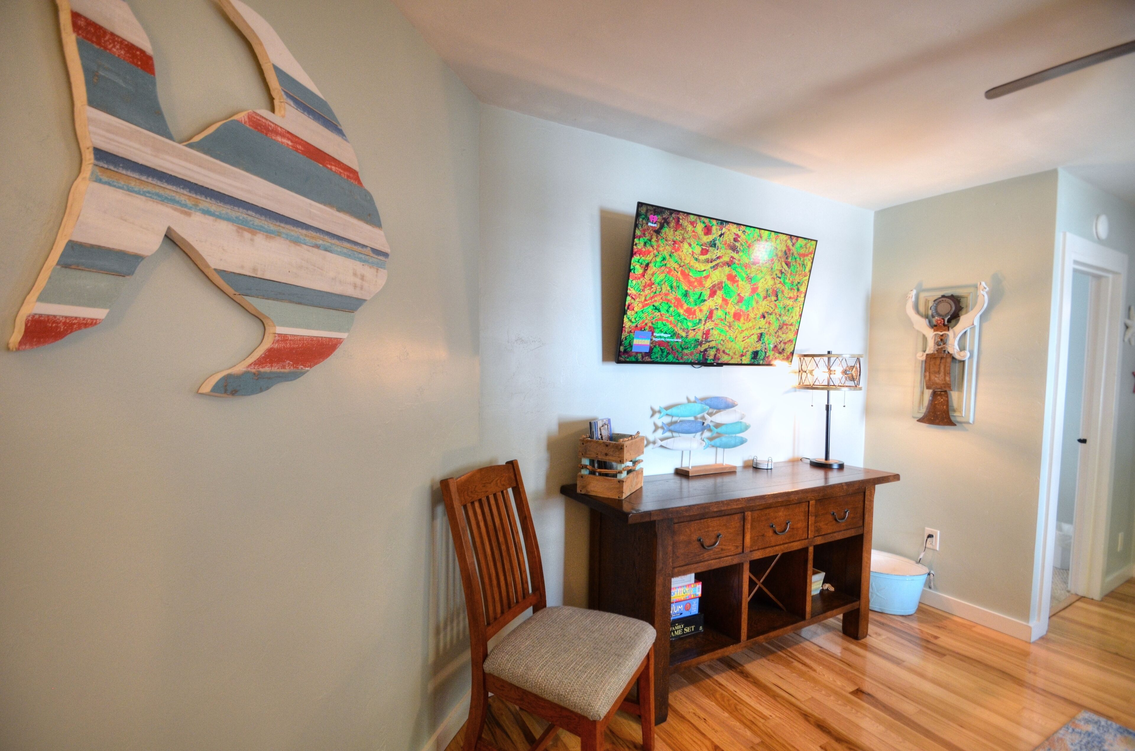 Une pièce comprenant une chaise en bois, un bureau en bois avec des tiroirs, une peinture abstraite aux couleurs vives et une sculpture de poisson accrochée au mur.