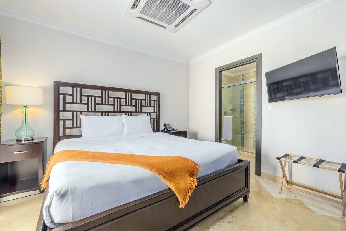 A neatly made bed with a patterned headboard, a bedside table with a lamp, and a TV mounted on the wall.