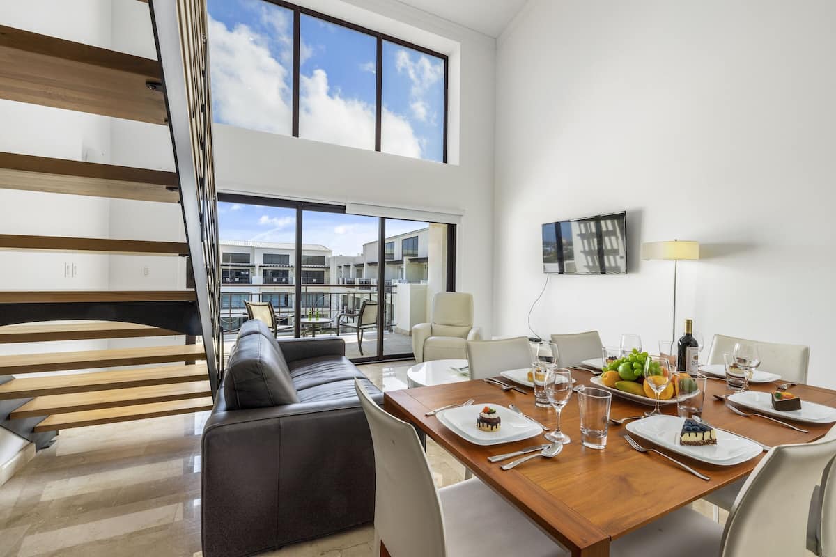 A modern dining area with a wooden table set for four, a black leather sofa, and a large window offering a view of the cityscape.