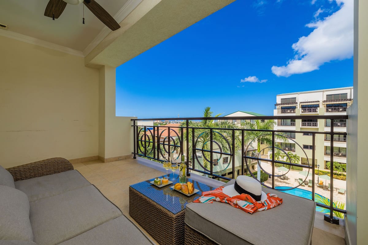A balcony with a view of a swimming pool and buildings, a wicker table with food and drinks, and a sofa.