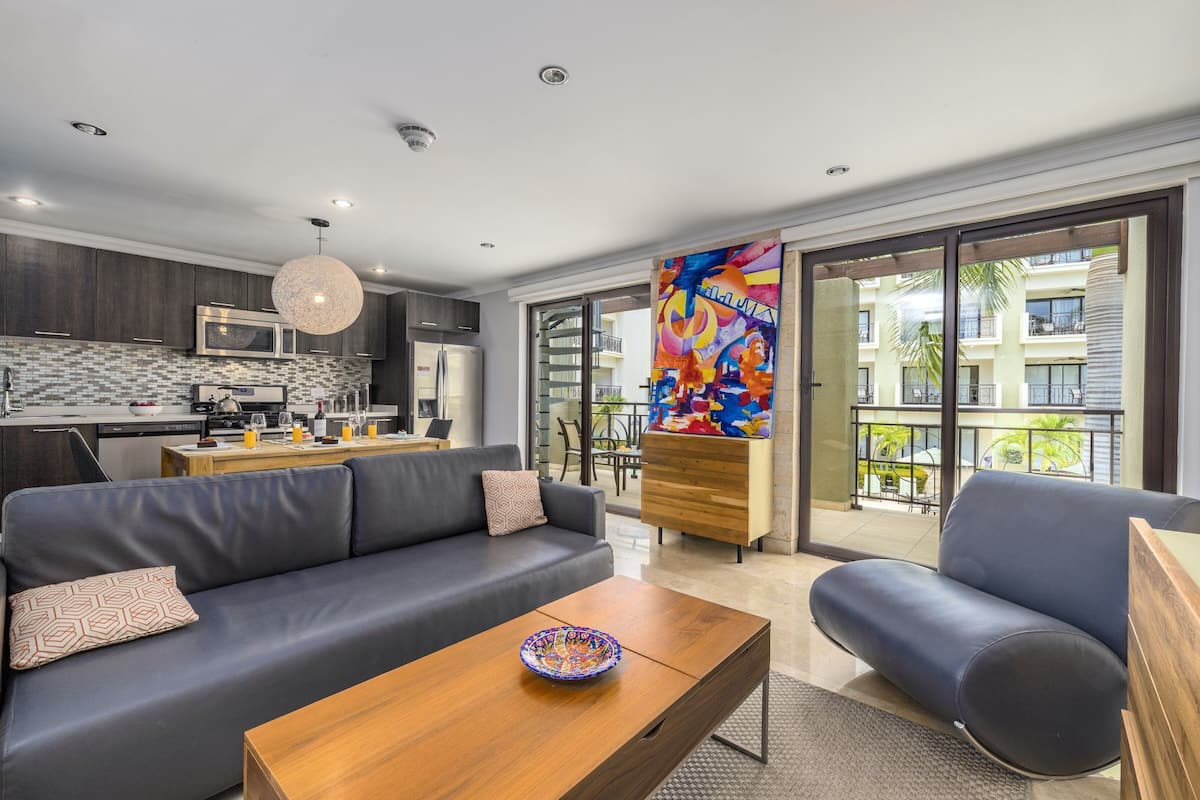 A modern living room with a sofa, armchair, and coffee table. A kitchen with dark wood cabinets and stainless steel appliances is visible in the background.