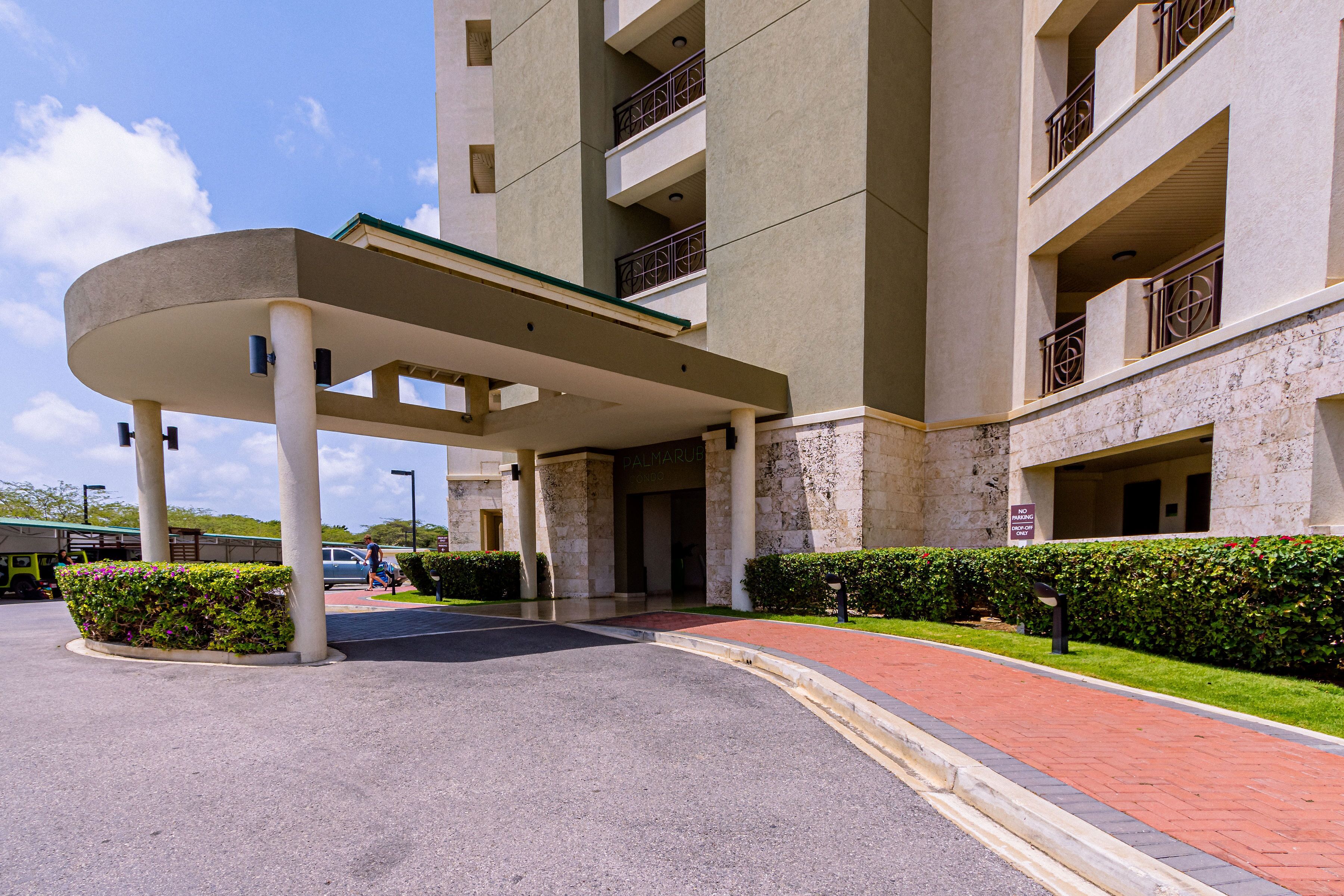 A modern building with a covered entrance, multiple balconies, and a landscaped area with hedges.