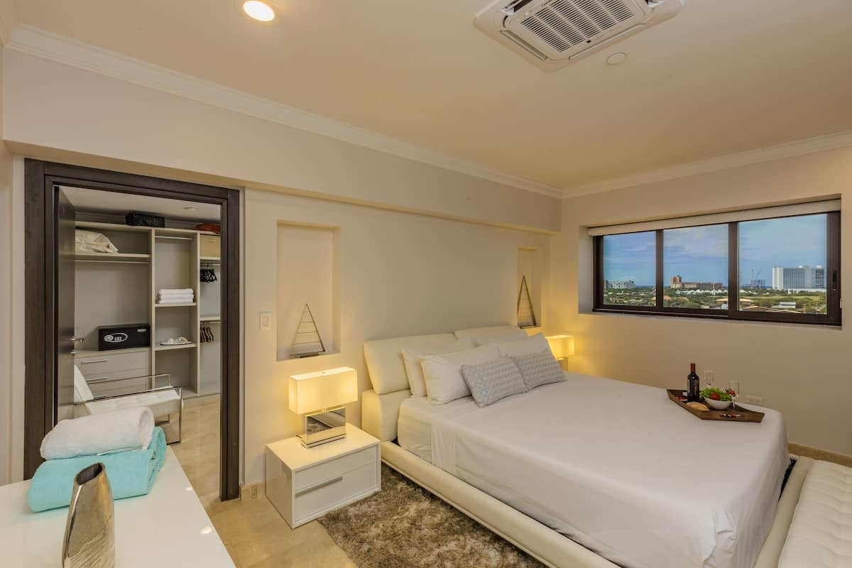 A bedroom with a bed, bedside tables, a nightstand, a window with a city view, and a closet with shelves and hanging items.