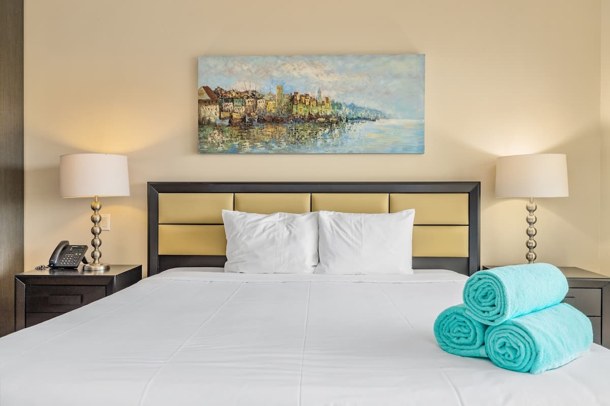 A neatly made bed with white linens, a headboard with a light wood finish, and two bedside lamps. A painting of a coastal town hangs above the bed.