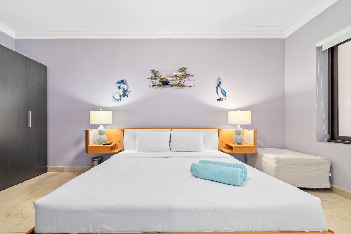 A hotel room with a large bed, two bedside tables with lamps, a small bedside table with a lamp, a nightstand, a window with a view, and a wall with decorative wall art.