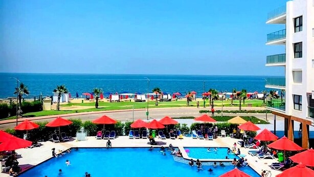 Área da piscina com guarda-sóis vermelhos, vista para o oceano e um edifício com varandas.