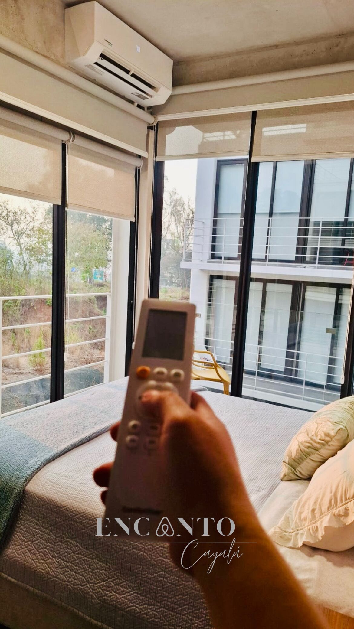 A hand holding a remote control in a modern hotel room with large windows, a bed, and an air conditioning unit.