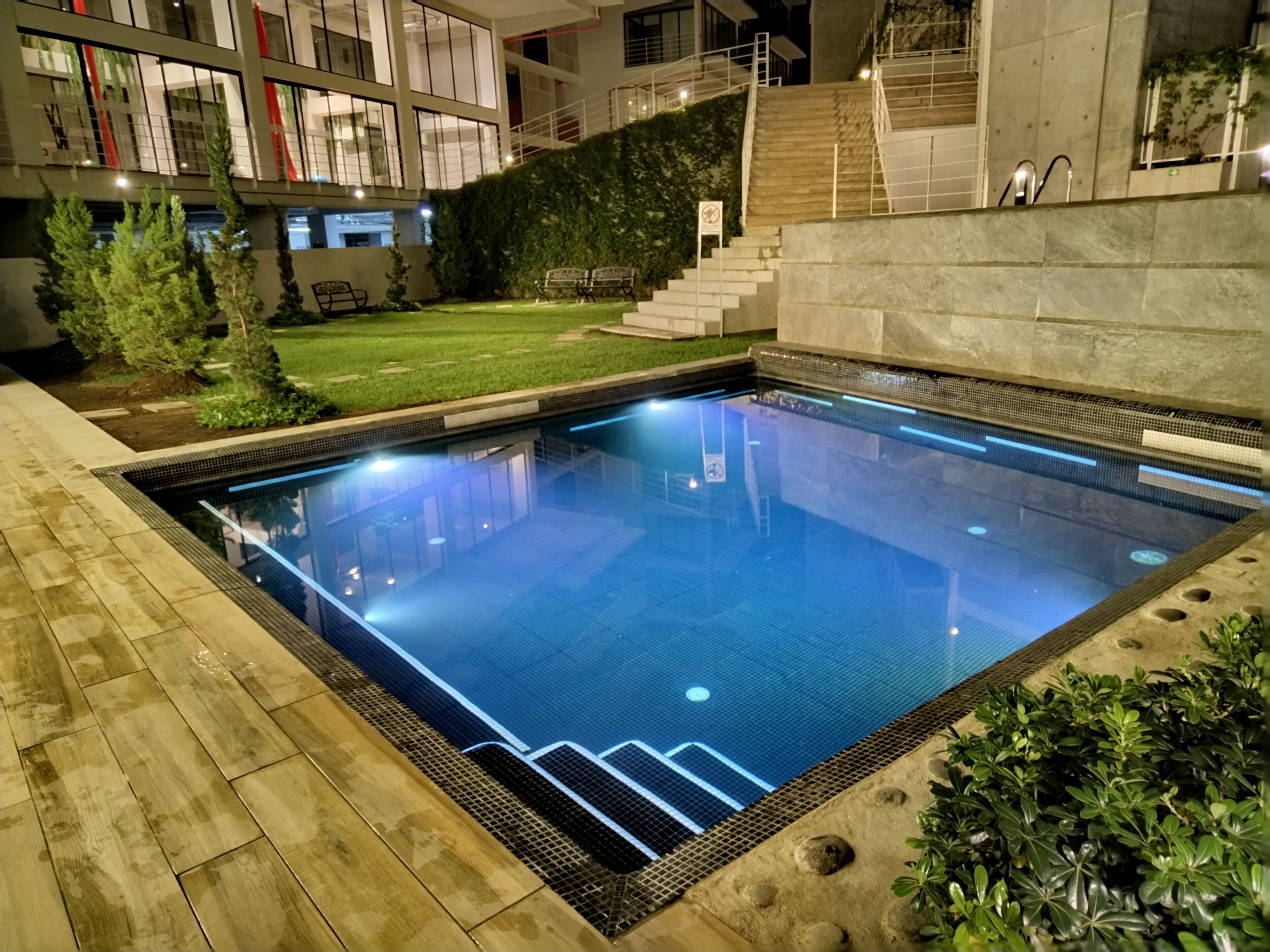 Una piscina con borde de baldosas, rodeada por un muro de piedra y una zona ajardinada con escaleras que suben a un edificio.