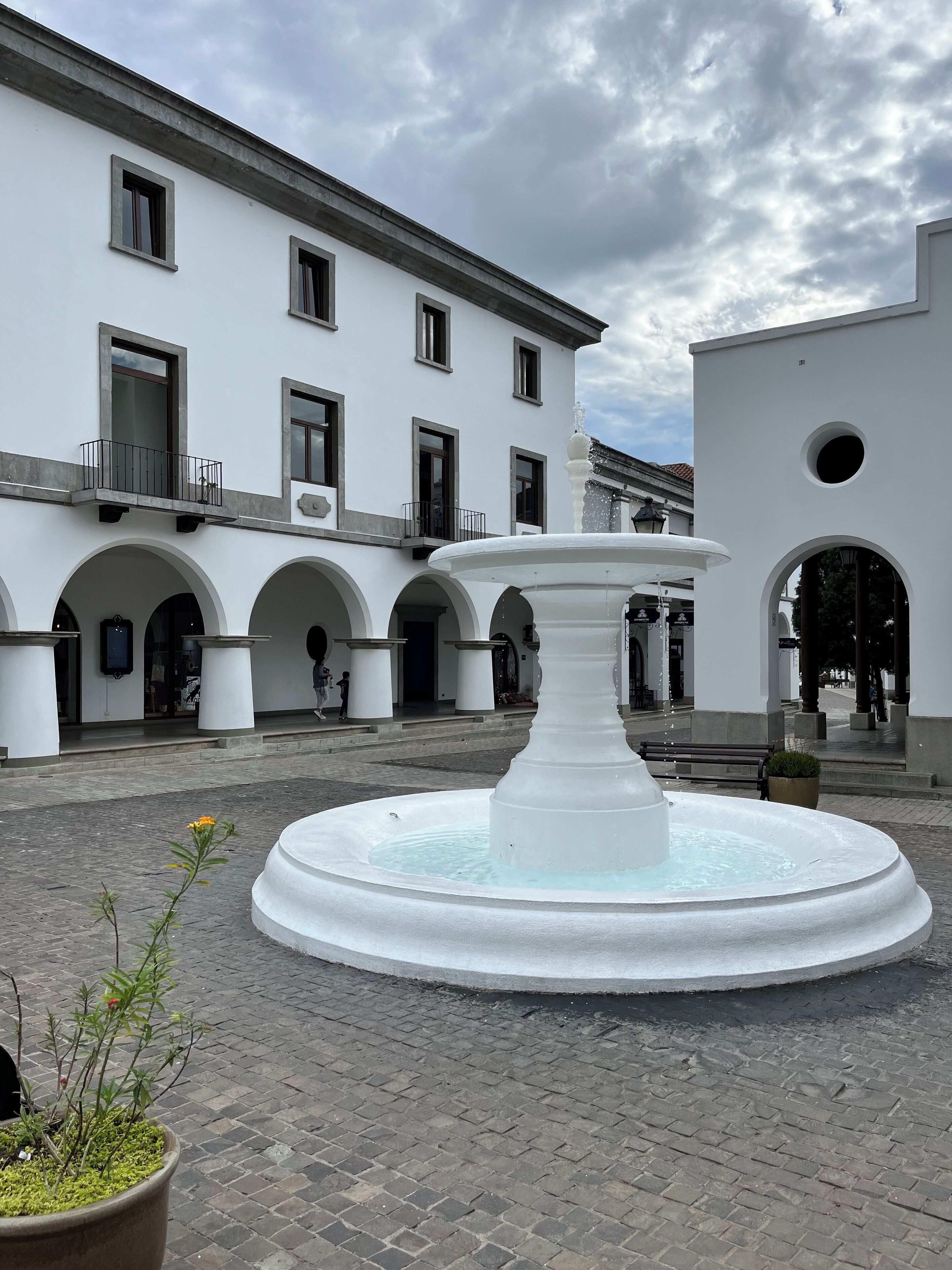 A courtyard with a central fountain, white buildings with arched doorways, and a paved ground.