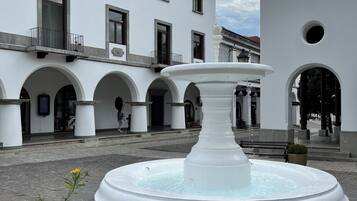 A courtyard with a central fountain, white buildings with arched doorways, and a paved ground.