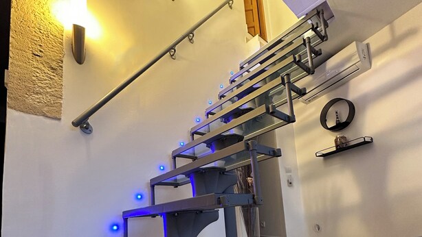 A modern staircase with glass steps and blue LED lighting, a round mirror, and a small shelf with a decorative item. - Lélégance de la Pierre (Fourques)