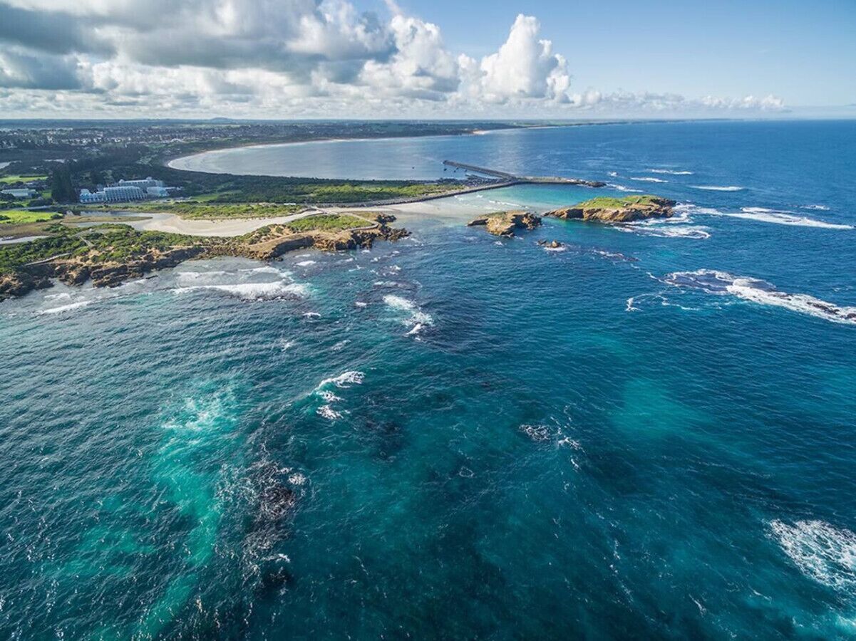An aerial view of a coastal area with clear blue waters, rocky outcrops, and a distant shoreline with buildings.