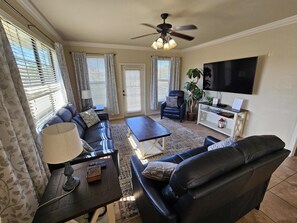 A living room with a sofa, coffee table, and armchairs, a TV mounted on the wall, and a ceiling fan.
