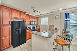 A kitchen with wooden cabinets, a black refrigerator, a microwave, a stove, and a granite countertop island with a chair. - Gorgeous, Spacious Condo With Balcony (Davenport)