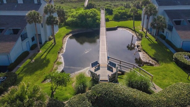 A coastal property with a central water feature, connected by a wooden bridge, surrounded by lush greenery and adjacent to a beach.