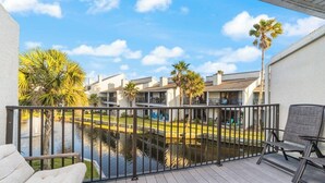 A balcony with a view of a water body and palm trees, wooden deck, and outdoor furniture.