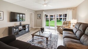 Ein Wohnzimmer mit Flachbildfernseher, einem dunkelbraunen Sofa, einem Glastisch und einem Deckenventilator.
