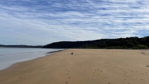 Une vaste plage de sable où l’on peut voir des empreintes de pas menant à l’eau, un ciel dégagé et, au loin, une colline boisée.