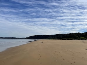 A wide sandy beach with footprints leading to the water, a clear sky, and a forested hill in the background. - Beachfront Family Holiday Home (Long Beach)