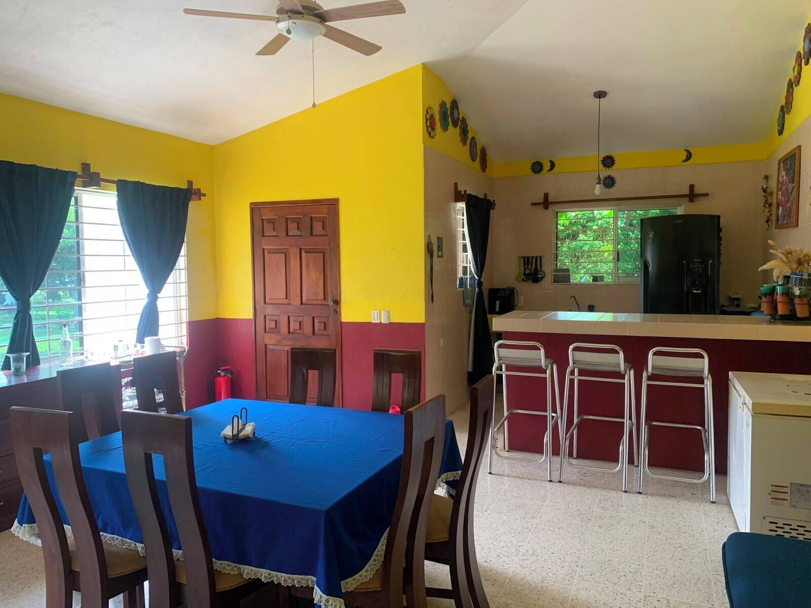 A dining area with a table set for four, a kitchen with a refrigerator, and a ceiling fan.