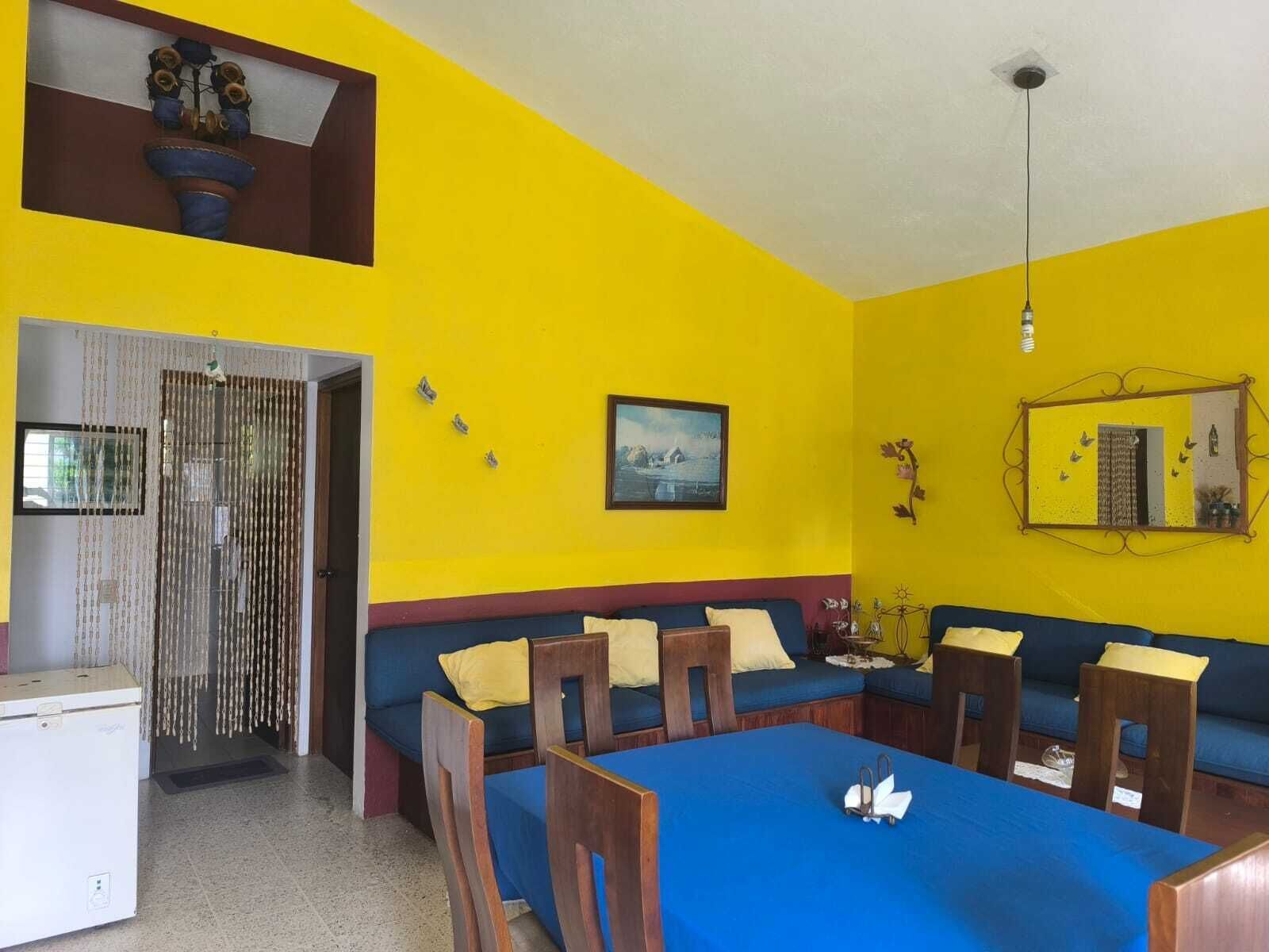 A room with a blue table, wooden chairs, a sofa, a small refrigerator, a decorative mirror, and a painting on the wall.