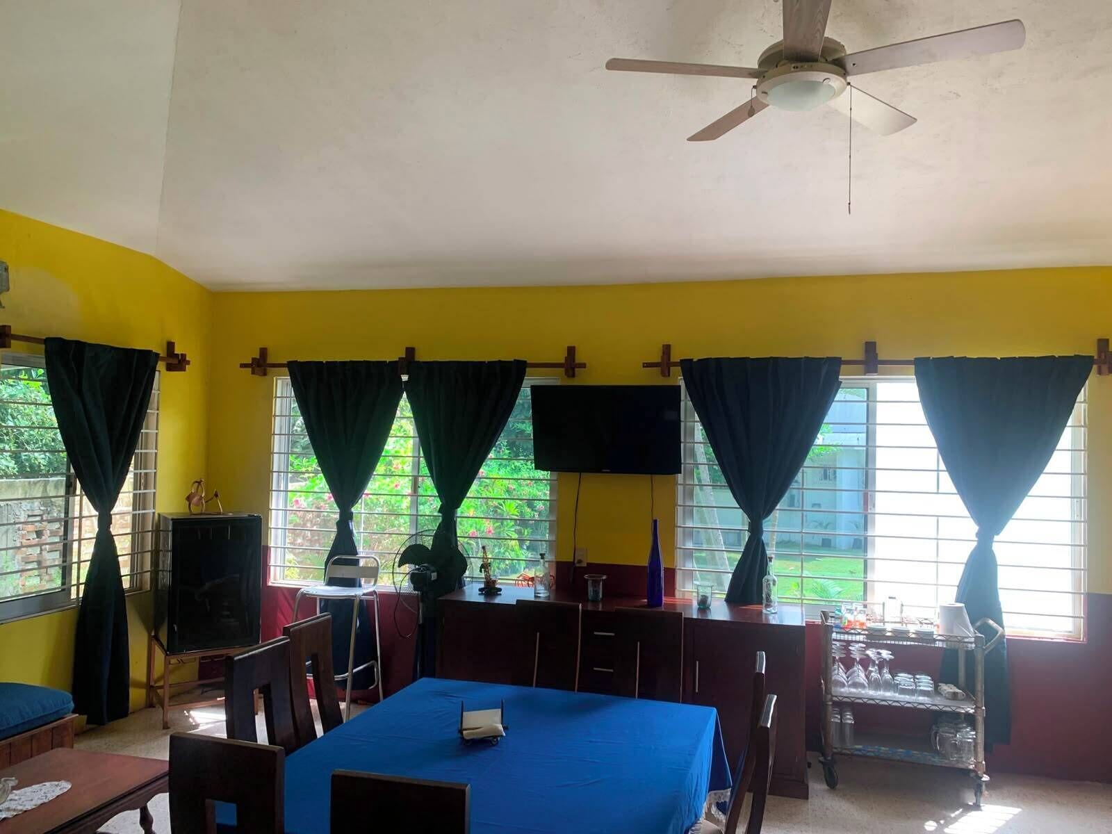 A room with a blue table, chairs, a ceiling fan, and windows with curtains.