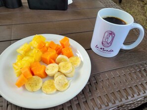 Une assiette blanche contenant des morceaux d’ananas, des dés d’orange et des tranches de banane, ainsi qu’une tasse blanche ornée d’un logo, le tout posé sur une table en bois.