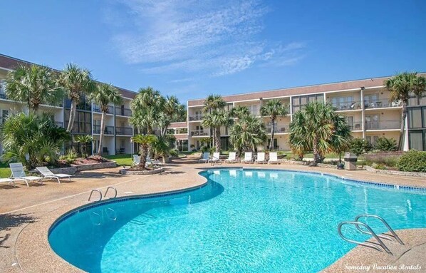 A swimming pool with lounge chairs and palm trees, surrounded by a multi-story building with balconies.