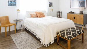 Un dormitorio con cama, una silla, una mesita, un armario y una ventana con cortinas.