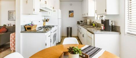A compact kitchen with white cabinets, a stove, microwave, and a sink. There is a dining table with a plant and a tray.