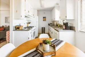 Una cocina compacta con gabinetes blancos, estufa, microondas y lavamanos. Hay una mesa de comedor con una planta y una bandeja.