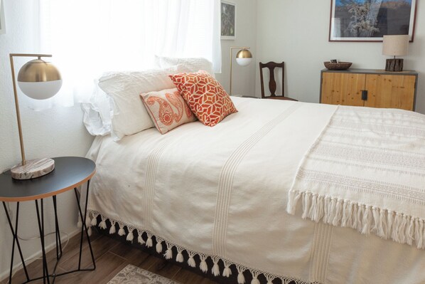 A neatly made bed with a white duvet and patterned pillows, a small round table with a lamp, and a wooden cabinet with a bowl on top.