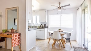 Una cocina moderna con armarios blancos, un comedor con mesa y sillas, un ventilador de techo y un amplio ventanal con persianas.