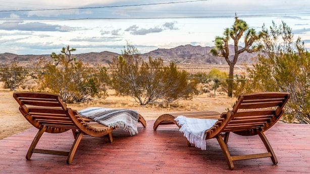 Dos sillones de madera con mantas sobre una terraza de madera con vistas a un paisaje desértico con montañas y árboles de Josué.