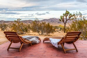 Two wooden lounge chairs with blankets on a wooden deck overlooking a desert landscape with mountains and Joshua trees.