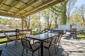 Une terrasse extérieure couverte, dotée d’une table et de chaises à plateau en verre, entourée d’arbres et offrant une vue sur un lac.
