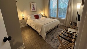 Un dormitorio con cama, ventilador de techo, ventana con persianas, mesita de noche con lámpara y alfombra.