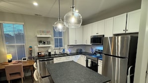 Una cocina moderna con una isla central, armarios blancos, electrodomésticos de acero inoxidable e iluminación colgante.