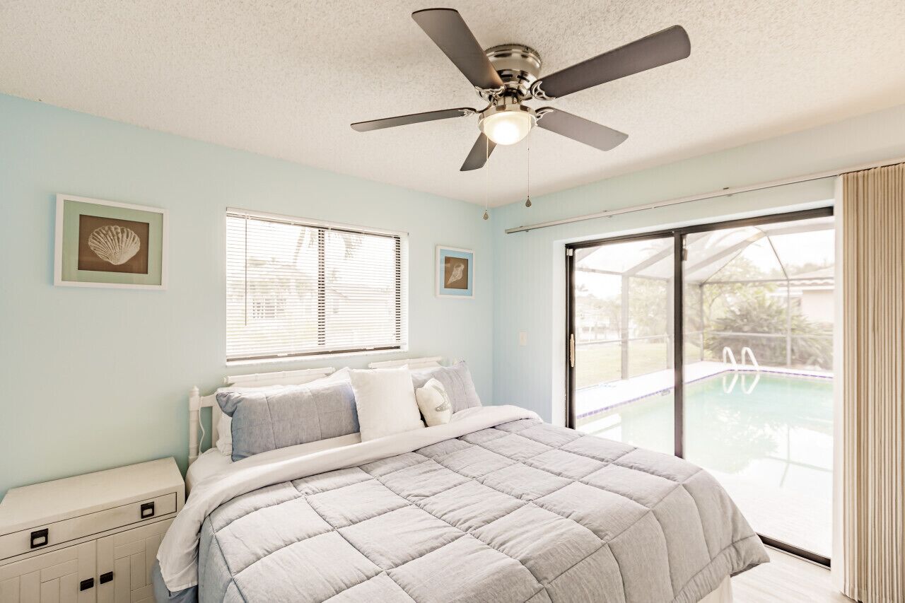 A bedroom with a bed, a ceiling fan, a window with blinds, and a view of an outdoor swimming pool.