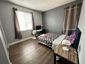 Cozy retreat with a single bed, desk, and chair, featuring a window with patterned curtains and a standing fan.