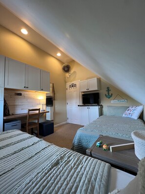Een compacte, goed verlichte kamer met een kitchenette, een bed met een 'Live Love Surf' kussen, een bureau met een gitaar en een kleine televisie.